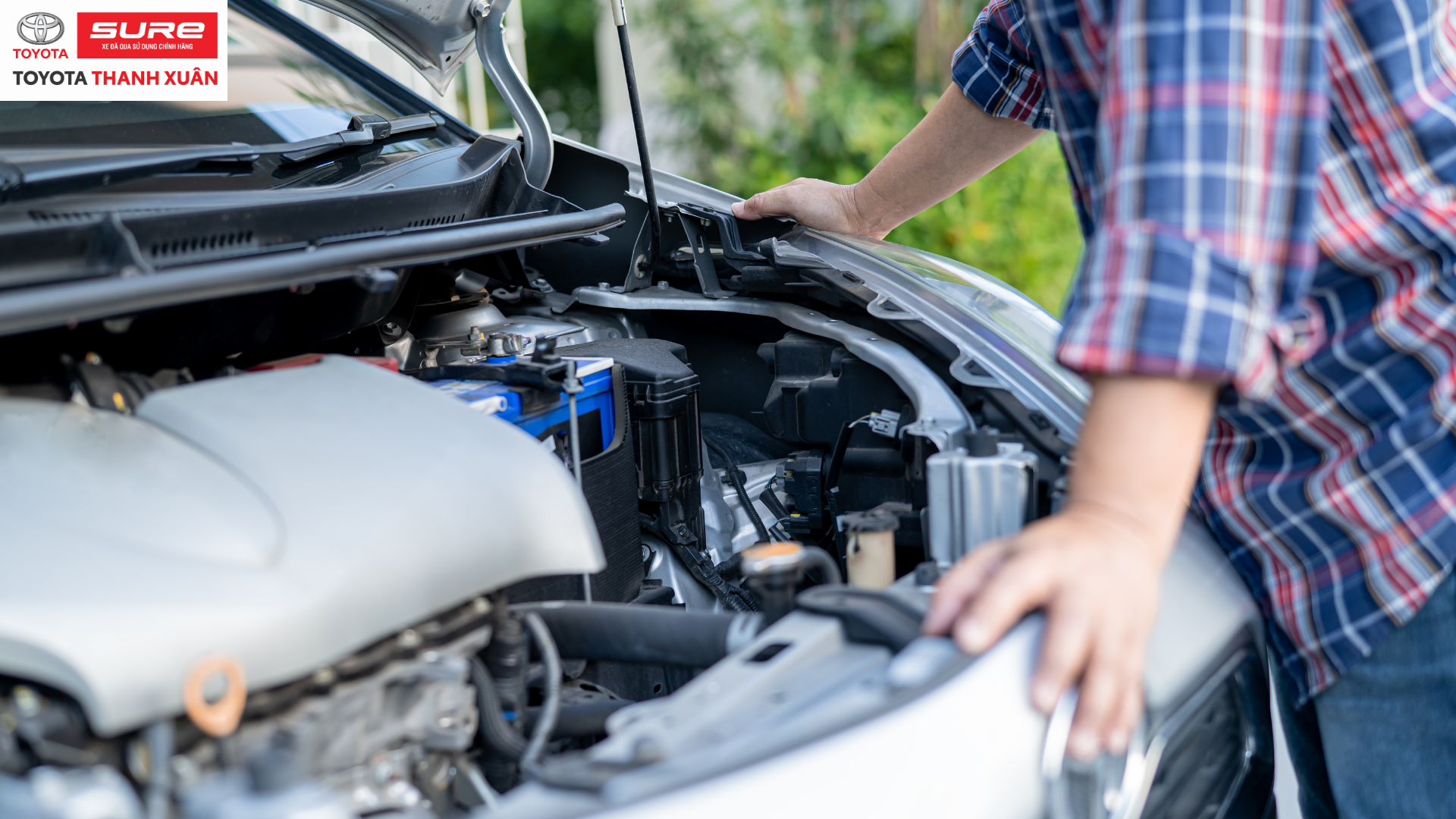 Người mặc áo kẻ kiểm tra động cơ xe hơi mở nắp, bối cảnh trời xanh, cây xanh. Trên góc trái có logo Toyota Sure Thanh Xuân.