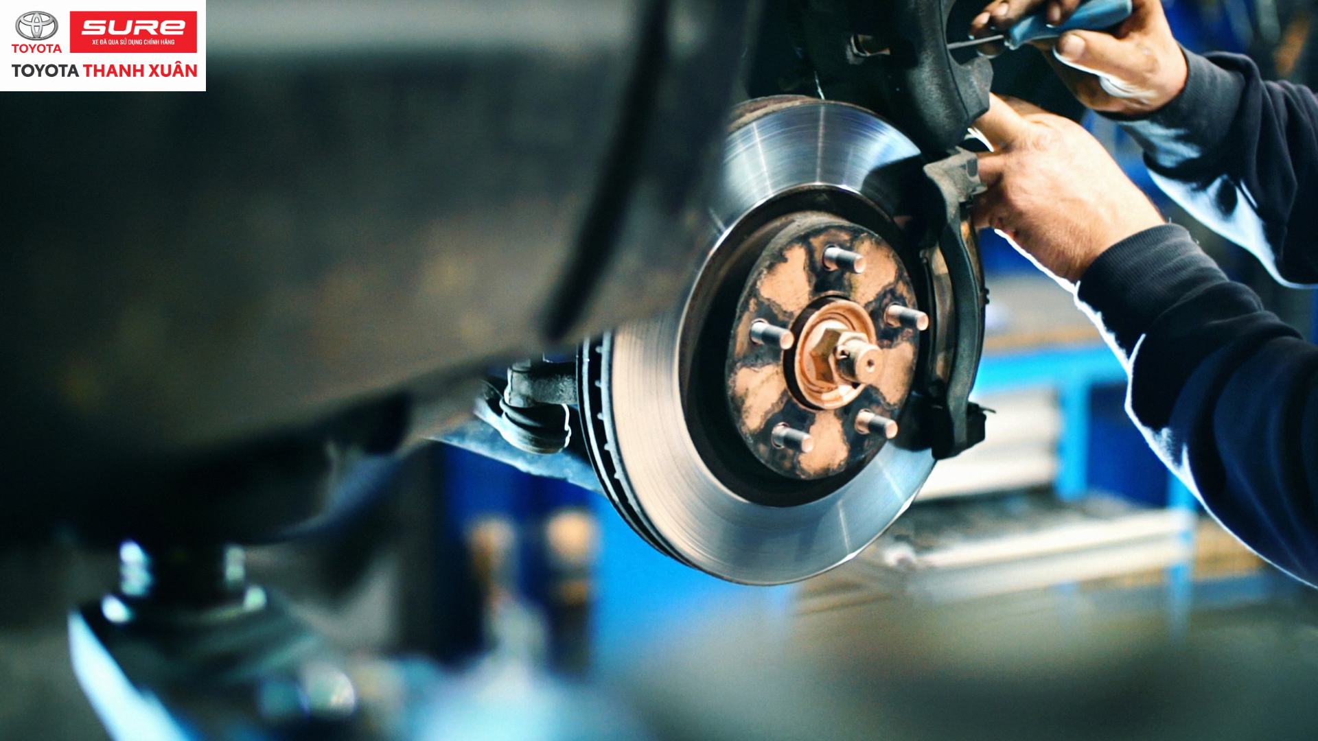 Kỹ thuật viên sửa phanh ô tô trong xưởng, tay áo tối màu, phanh kim loại sáng bóng. Logo Toyota Sure Thanh Xuân phía trên.